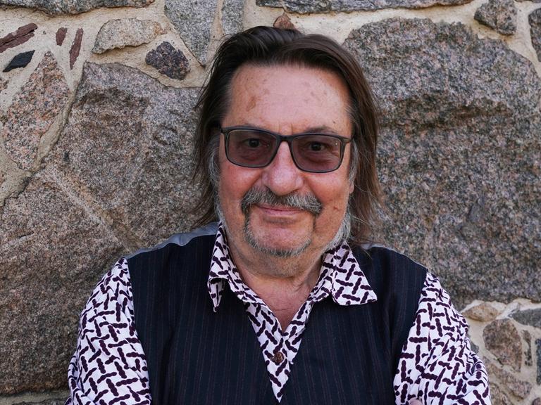 Ein Mann mit einer getönten Brille steht vor einer Steinwand. Er trägt kariertes Hemd und eine dunkle Weste. Er hält die Arme verschränkt und blickt freundlich. Es ist der Musiker Manfred Maurenbrecher.