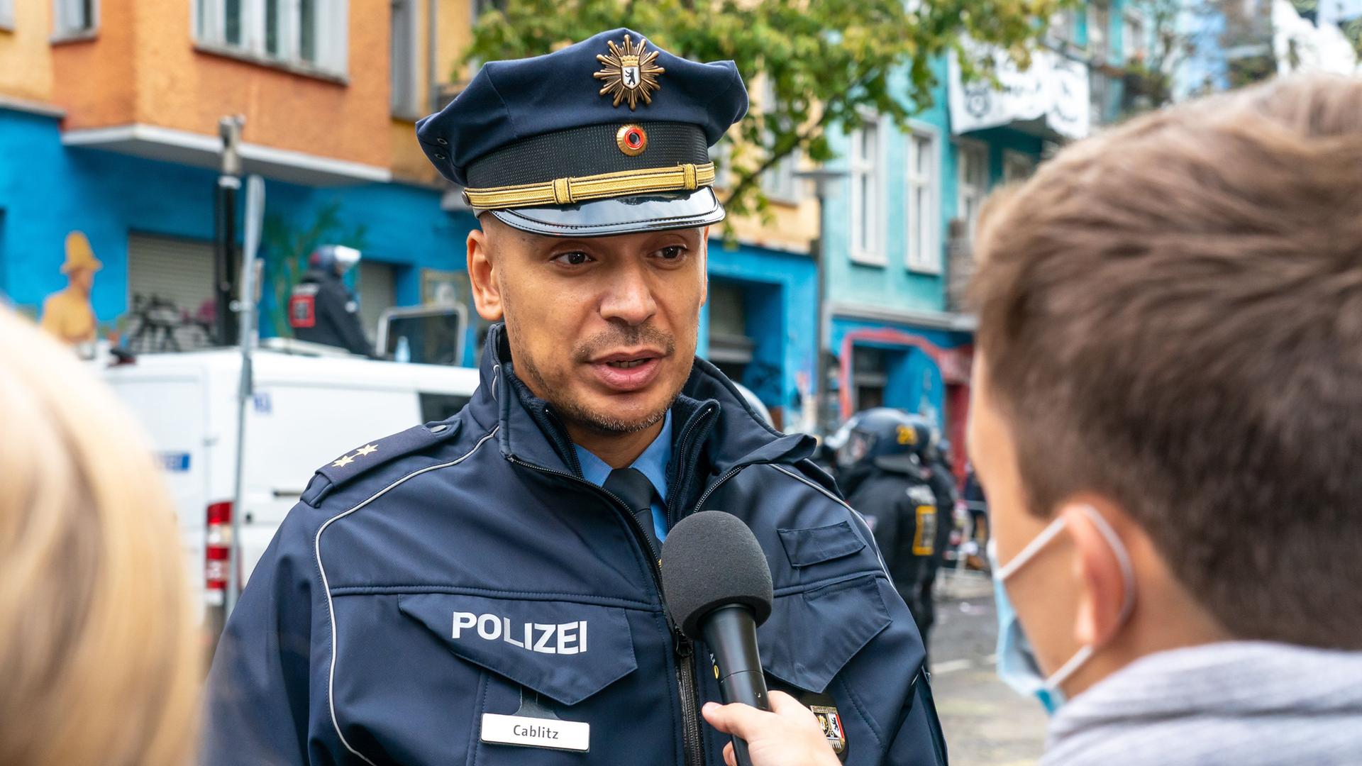 Thilo Calblitz in Polizeiuniform unf mit Polizeimütze auf dem Kopf spricht mit einem anderen Menschen, der ein Mikrofon in der Hand hält. Diese Person ist von hinten zu sehen. Thilo Calblitz in Polizeiuniform unf mit Polizeimütze auf dem Kopf spricht mit einem anderen Menschen, der ein Mikrofon in der Hand hält. Diese Person ist von hinten zu sehen.