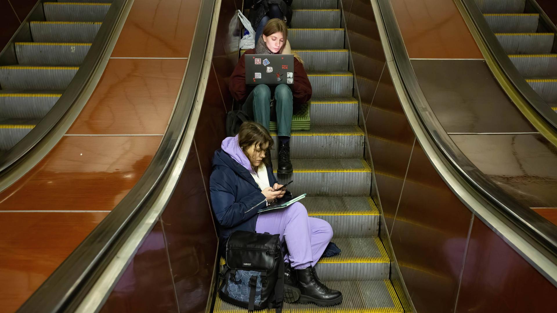 Junge Menschen mit Smartphone und Laptop sitzen auf einer Rolltreppe in einer U-Bahn Station in Kiew und warten einen Luftangriffalarm ab. 13. Dezember 2022, Kiew, Ukraine. Junge Menschen mit Smartphone und Laptop sitzen auf einer Rolltreppe in einer U-Bahn Station in Kiew und warten einen Luftangriffalarm ab. 13. Dezember 2022, Kiew, Ukraine.