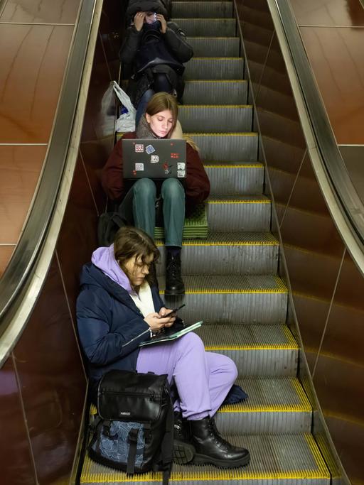 Junge Menschen mit Smartphone und Laptop sitzen auf einer Rolltreppe in einer U-Bahn Station in Kiew und warten einen Luftangriffalarm ab. 13. Dezember 2022, Kiew, Ukraine.