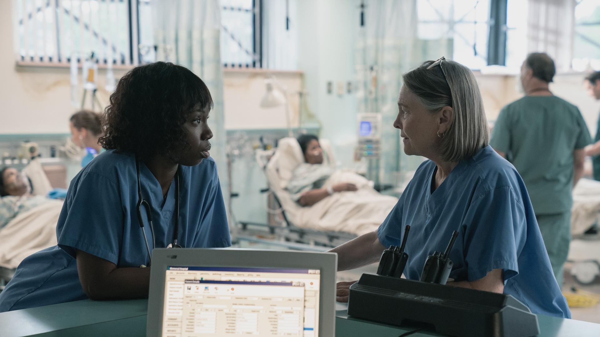 Zwei Frauen in stehen am Tresen einer Krankenhaus-Station. Im Hintergrund sind Patienten in Betten zu sehen.