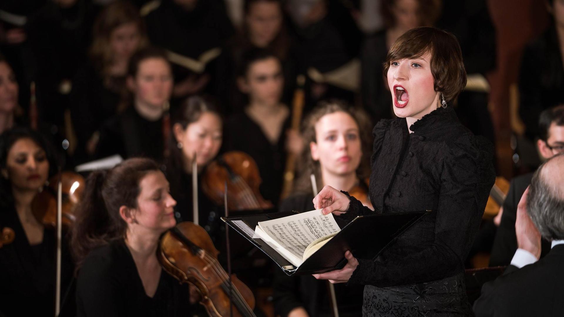 Eine Frau steht mit Noten in der Hand singend in einem Orchester Eine Frau steht mit Noten in der Hand singend in einem Orchester