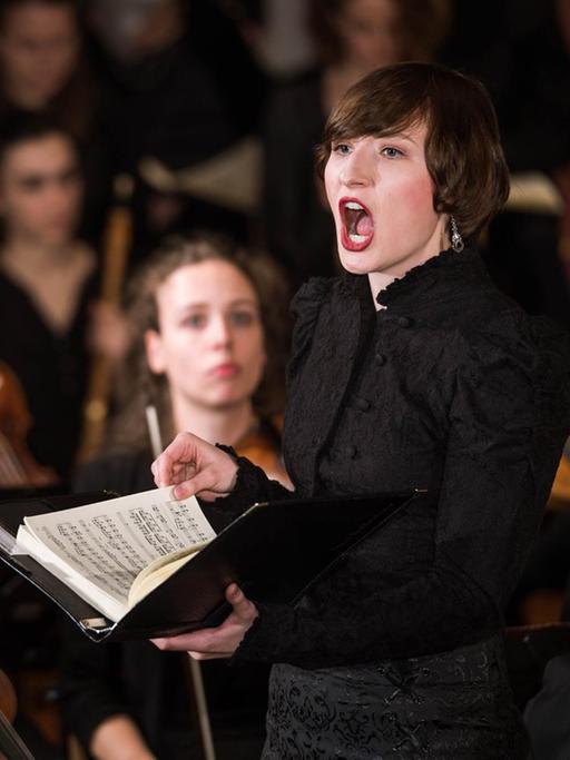 Eine Frau steht mit Noten in der Hand singend in einem Orchester