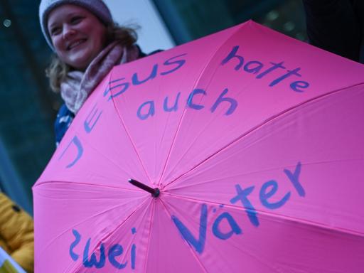 Eine Frau hält bei Protesten am Rande der Synodalversammlung einen Regenschirm mit der Aufschrift "Jesus hatte auch zwei Väter". 