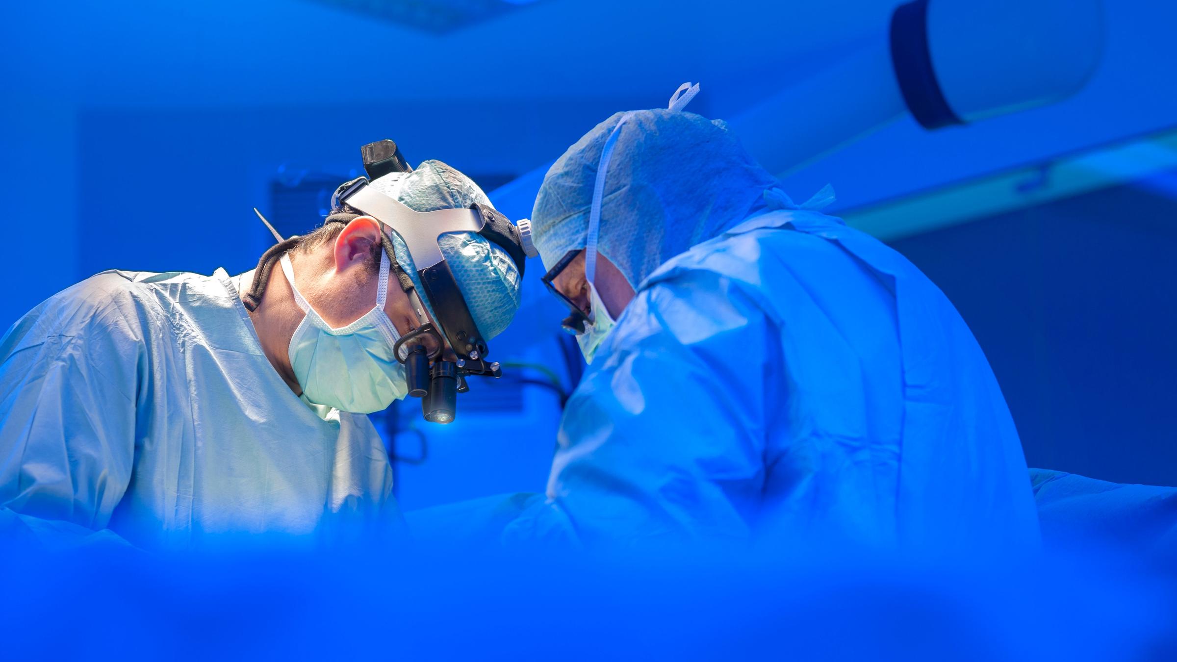 Zwei Ärzte in OP-Kleidung stehen in einem OP-Saal und scheinen zu operieren. Das Licht ist kühl, sie tragen helle Kleidung. 