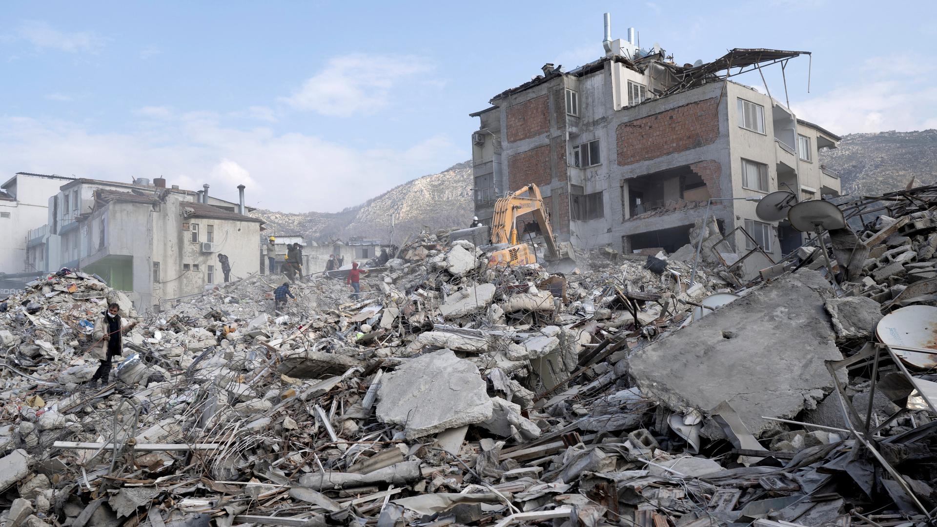 Mehrere Menschen klettern in Hatay über die Trümmer eingestürzter Häuser. Auf dem Schuttberg steht auch ein Bagger.