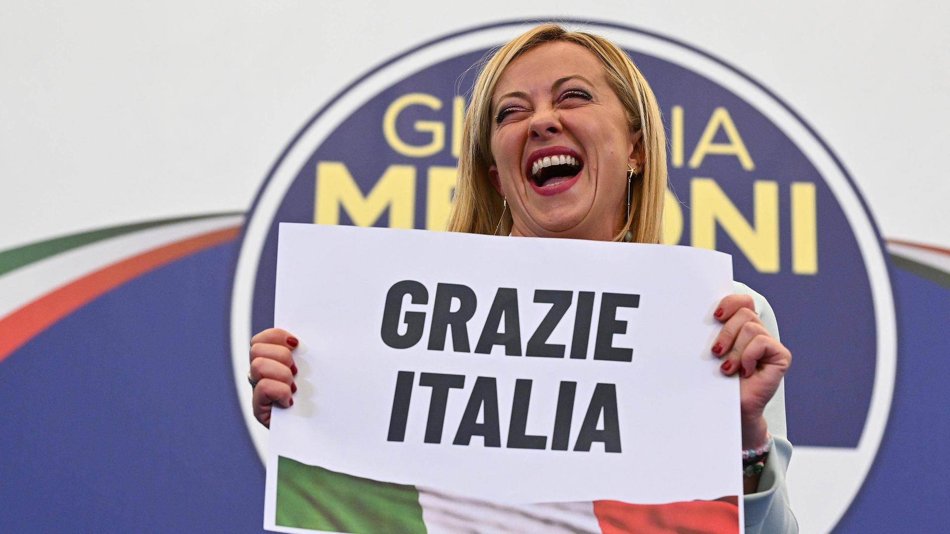 Meloni steht auf einer Bühne mit dem Logo ihrer Partei, lacht und hält ein Schild mit der Aufschrift "Grazie Italia" in der Hand. 
