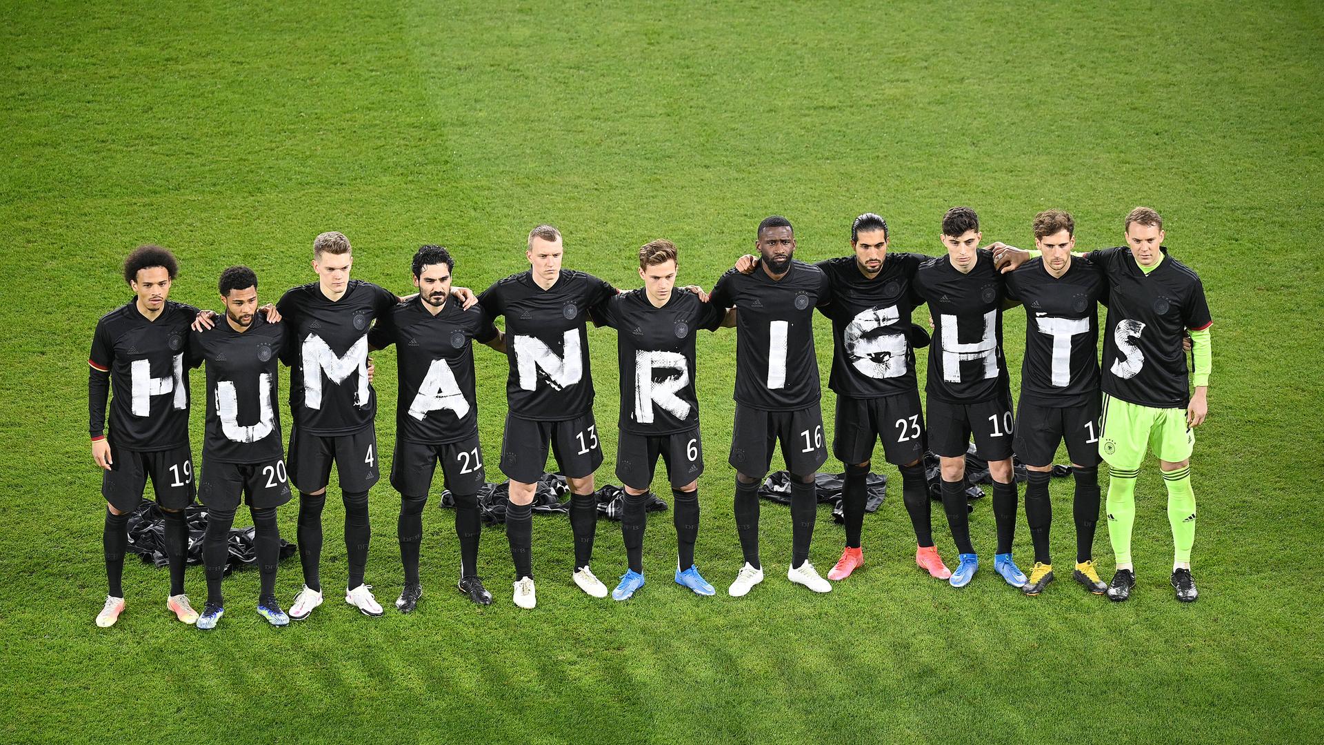 Fußballer stehen auf dem Rasen in einer Reihe und haben jeweils ein schwarzes T-Shirt an, auf dem mit weißer Farbe Human Rights geschrieben steht. Fußballer stehen auf dem Rasen in einer Reihe und haben jeweils ein schwarzes T-Shirt an, auf dem mit weißer Farbe Human Rights geschrieben steht.