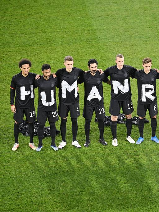Fußballer stehen auf dem Rasen in einer Reihe und haben jeweils ein schwarzes T-Shirt an, auf dem mit weißer Farbe Human Rights geschrieben steht.
