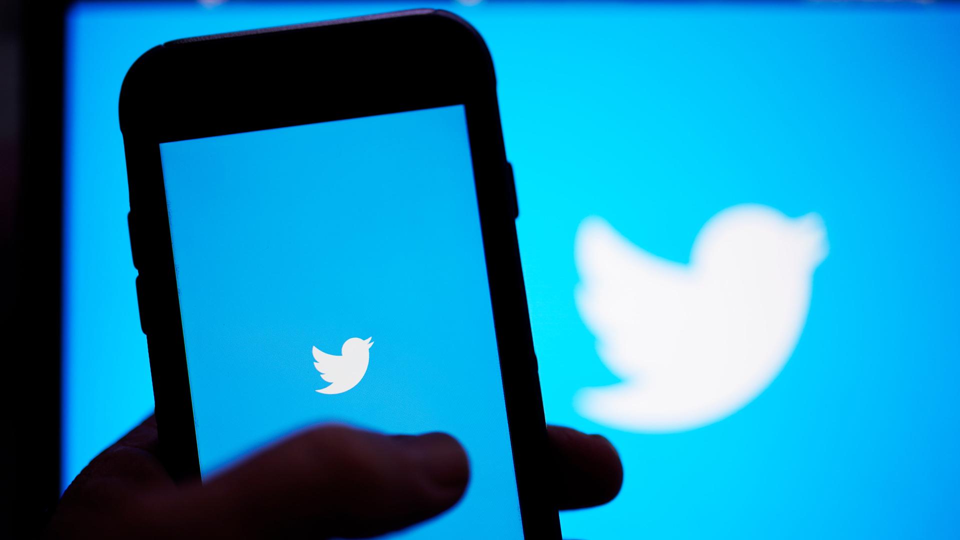 Ein Hand hält ein Smartphone auf dessen Bildschirm das Twitter-Symbol zu sehen ist, das auch im Hintergrund erscheint Ein Hand hält ein Smartphone auf dessen Bildschirm das Twitter-Symbol zu sehen ist, das auch im Hintergrund erscheint