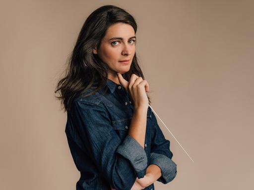 Eine junge Frau mit braunen langen Haaren hat die Arme halb verschränkt. Den Kopf stützt sie auf den Zeigefinger ihrer rechten Hand. In der Hand hält sie auch einen Taktstock. Ihr Blick ist freundlich.