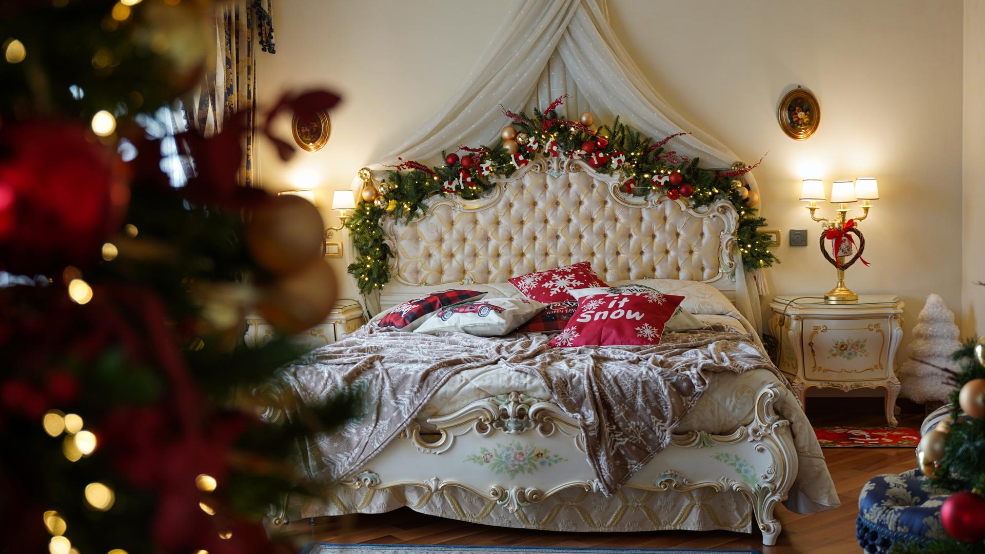 Ein barockes Bett ist mit festlicher Weihnachtsdekoration am Bettgestell geschmückt. Auf der Decke liegen Kissen mit Christmas-Beschriftungen.