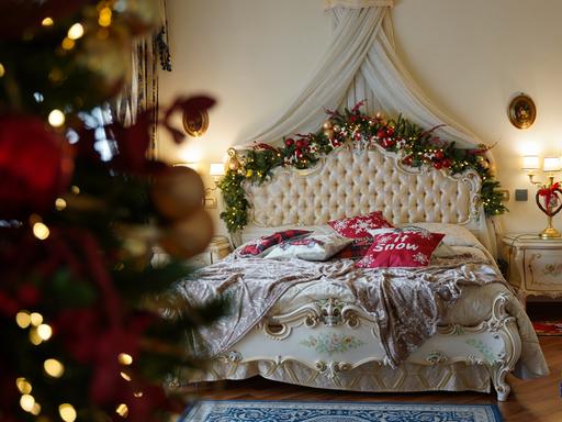 Ein barockes Bett ist mit festlicher Weihnachtsdekoration am Bettgestell geschmückt. Auf der Decke liegen Kissen mit Christmas-Beschriftungen.