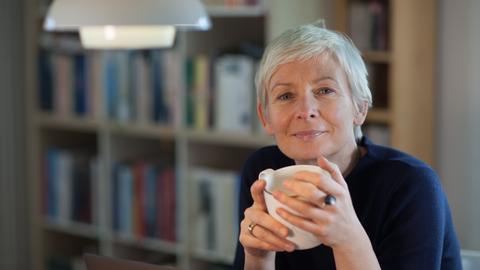 Portrait von Dörte Hansen, sie hät eine Tasse in beiden Händen, im HIntergrund ein Bücherregal