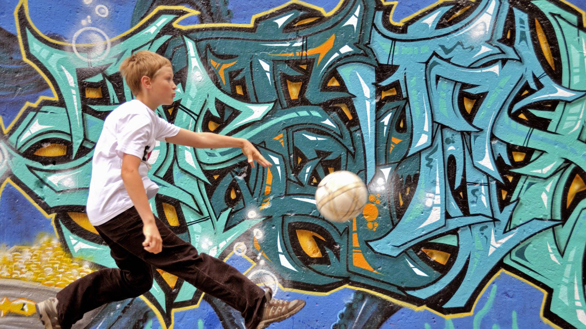 Junge spielt alleine Fussball auf der Strasse vor einer Graffitiwand
