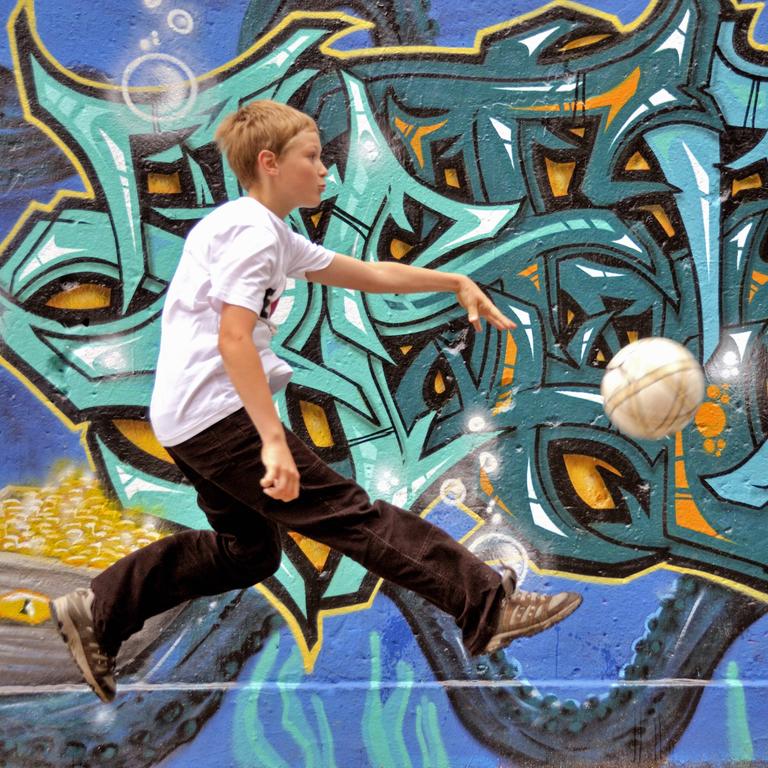 Boy playing soccer alone on the street in front of a graffiti wall