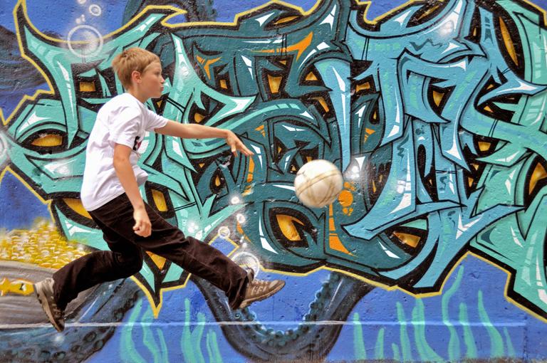 Junge spielt alleine Fussball auf der Strasse vor einer Graffitiwand
