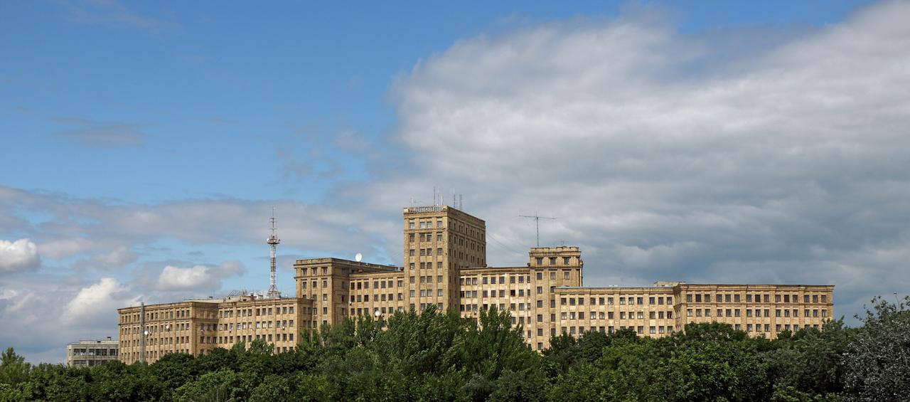 Das langgestreckte Universitätsgebäude von Charkiw erhebt sich über einem grünen Laubwald vor strahlend blauem Himmel.