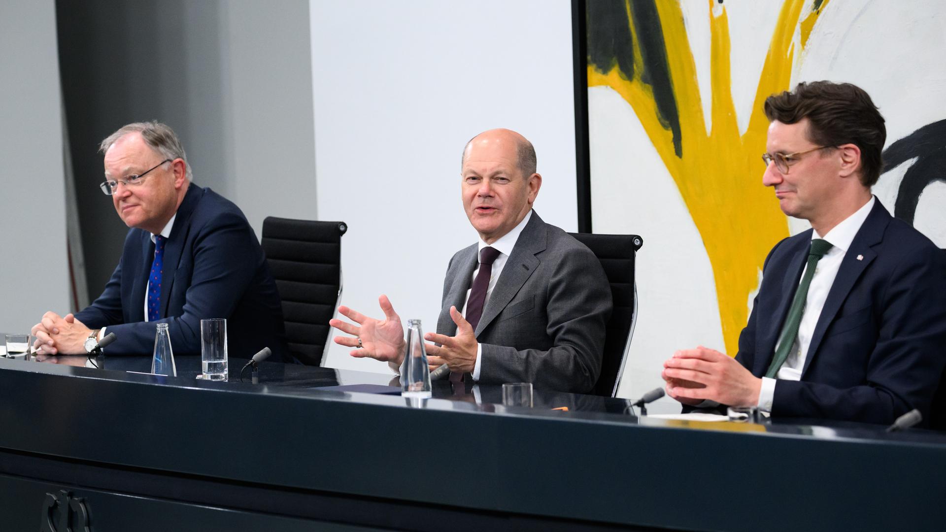 Weil, Scholz und Wüst sitzen bei der Pressekonferenz hinter einem langen Tisch. Im Hintergrund ist ein großes, buntes Wandgemälde zu sehen. 