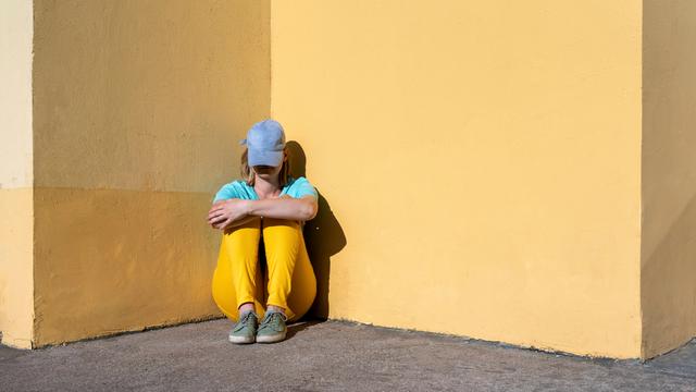 Eine junge Frau sitzt mit ins Gesicht gezogener Baseballcap, an einer Wand gelehnt auf dem Boden.