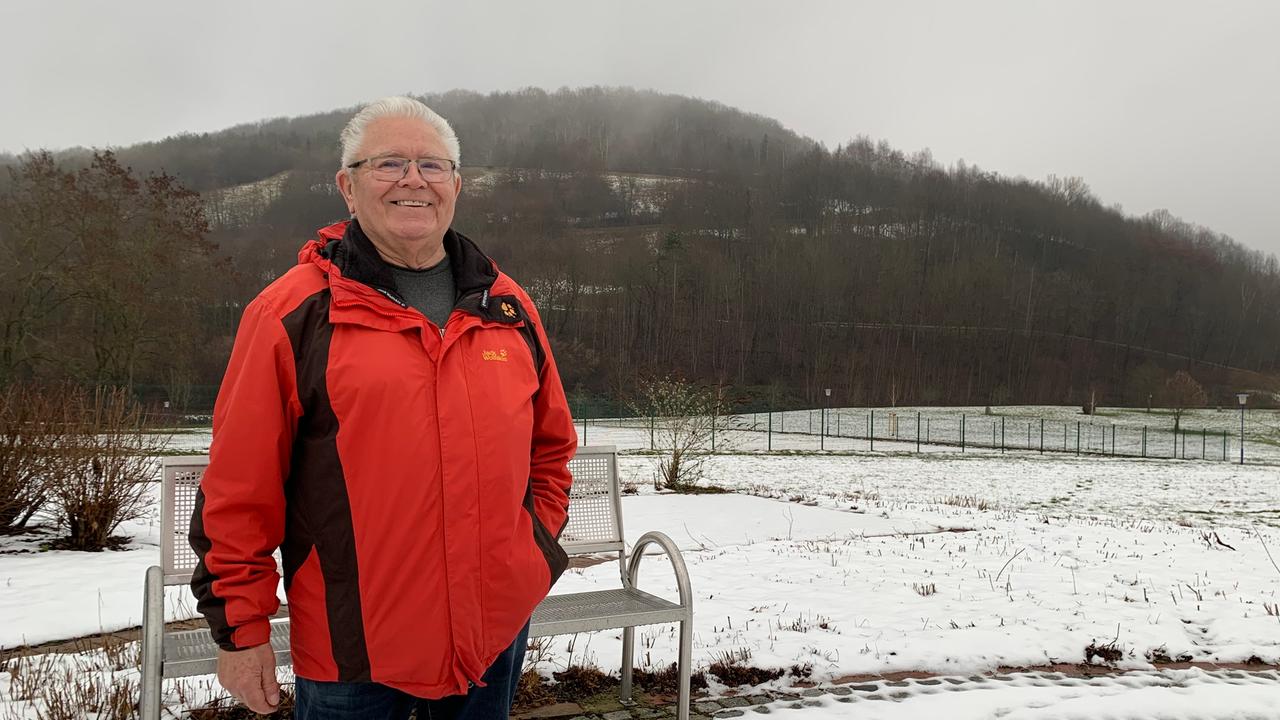 Adolf Vater ist Anfang 70, graue Haare, rote Jacke. Er steht vor einem schneebedeckten, bewaldeten Hügel.