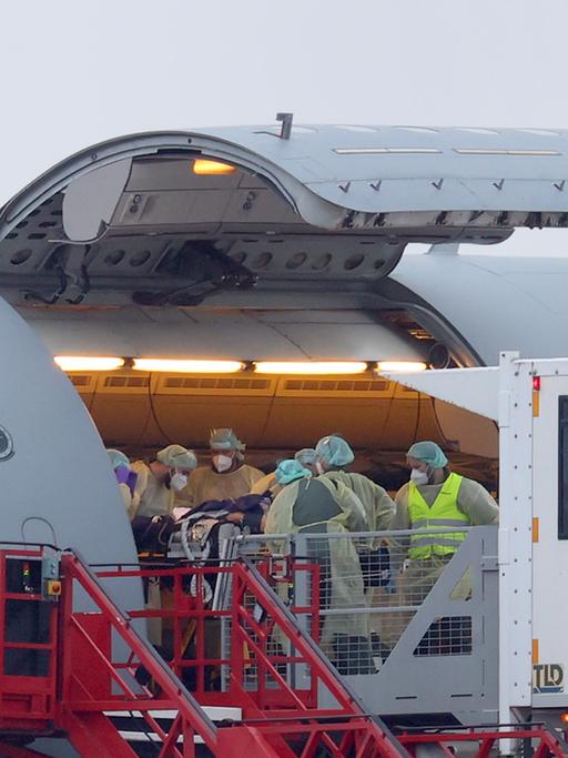 Mehrere Helfer in medizinischer Schutzkleidung schieben einen schwer kranken Patienten auf einer Liege in ein Flugzeug.