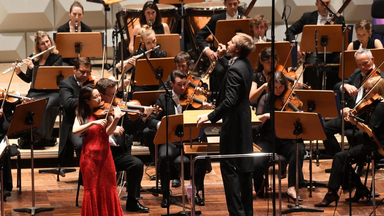 Geigerin Bomsori Kim trägt ein rotes Abendkleid und spielt ihre Geige. Im Hintergrund das Royal Philharmonic Orchestra. Geigerin Bomsori Kim trägt ein rotes Abendkleid und spielt ihre Geige. Im Hintergrund das Royal Philharmonic Orchestra.