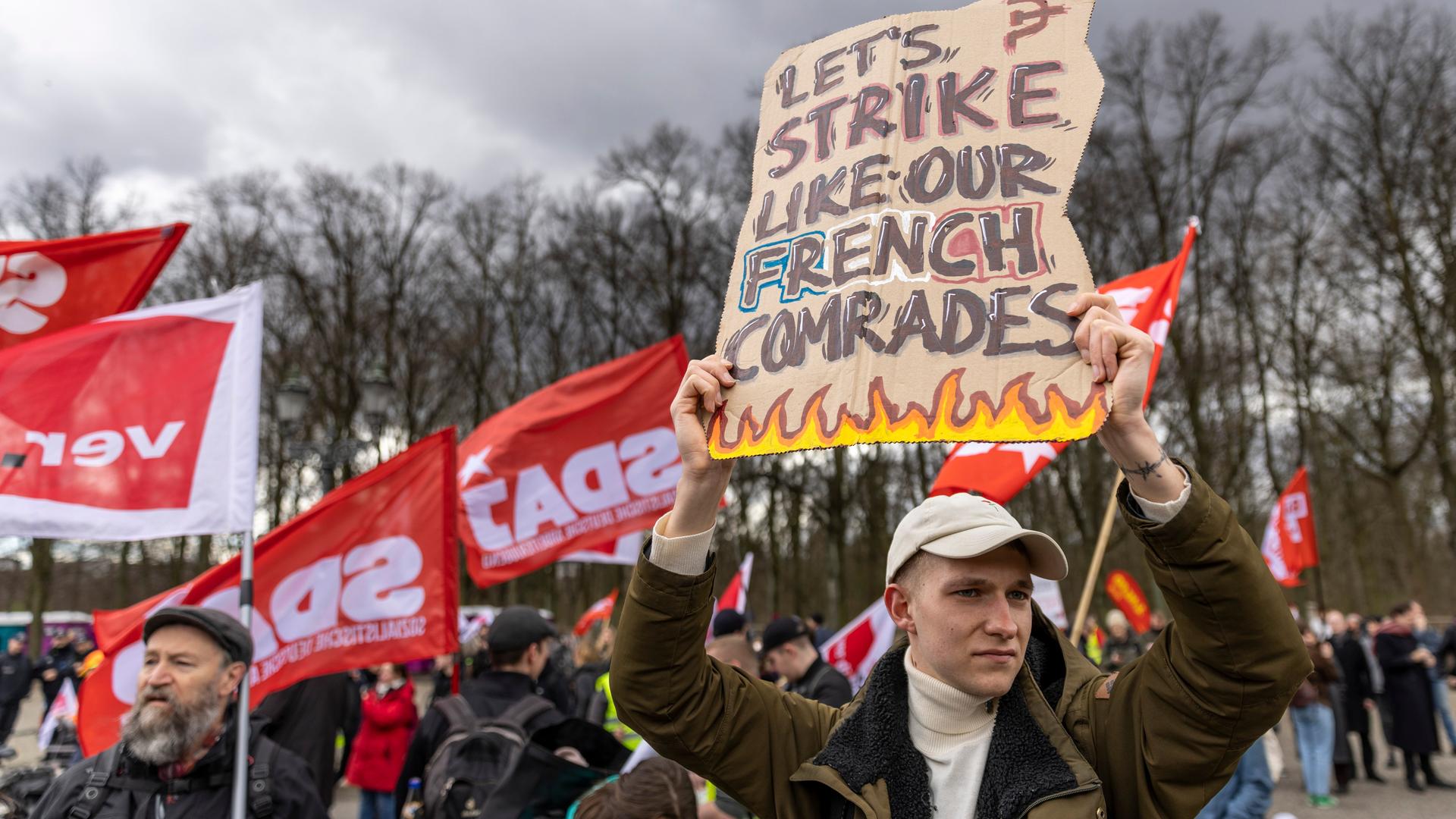 Eine ganze Reihe Personen nehmen an einem Gewerkschaftsprotest am 25. März 2023 teil. Ein Mann hält ein Schild hoch, auf dem steht "Let's strike like out French comrades". Eine ganze Reihe Personen nehmen an einem Gewerkschaftsprotest am 25. März 2023 teil. Ein Mann hält ein Schild hoch, auf dem steht "Let's strike like out French comrades".
