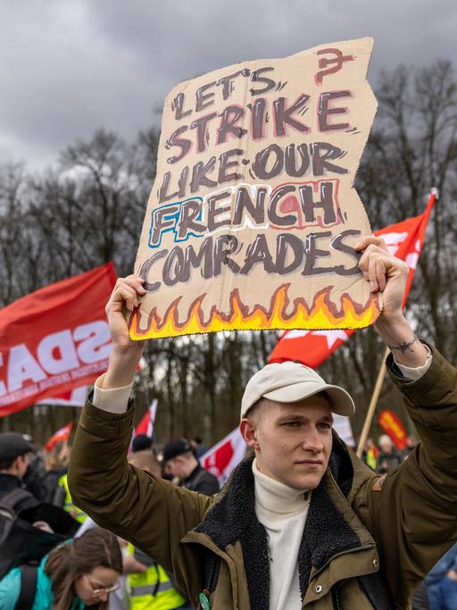 Eine ganze Reihe Personen nehmen an einem Gewerkschaftsprotest am 25. März 2023 teil. Ein Mann hält ein Schild hoch, auf dem steht "Let's strike like out French comrades".