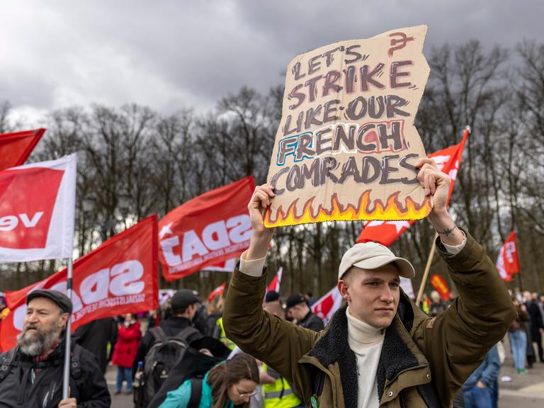 Eine ganze Reihe Personen nehmen an einem Gewerkschaftsprotest am 25. März 2023 teil. Ein Mann hält ein Schild hoch, auf dem steht "Let's strike like out French comrades".