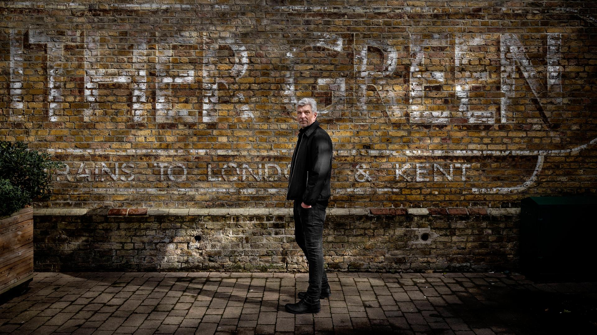 Ein Mann mit kurzen grauen Haaren steht vor einer alten Backsteinmauer. Ein Mann mit kurzen grauen Haaren steht vor einer alten Backsteinmauer.