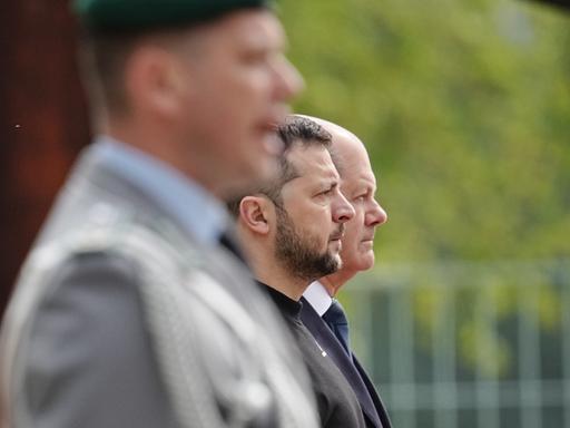 Zwei Männer im Profil beobachten mit strengem Blick eine Militärparade außerhalb des Blickfelds, ein unscharfer Militär im Vordergrund gibt Kommandos