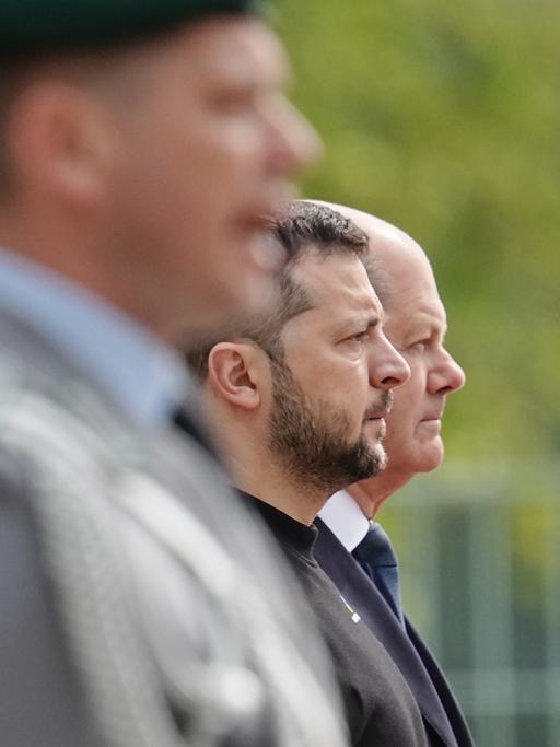 Zwei Männer im Profil beobachten mit strengem Blick eine Militärparade außerhalb des Blickfelds, ein unscharfer Militär im Vordergrund gibt Kommandos
