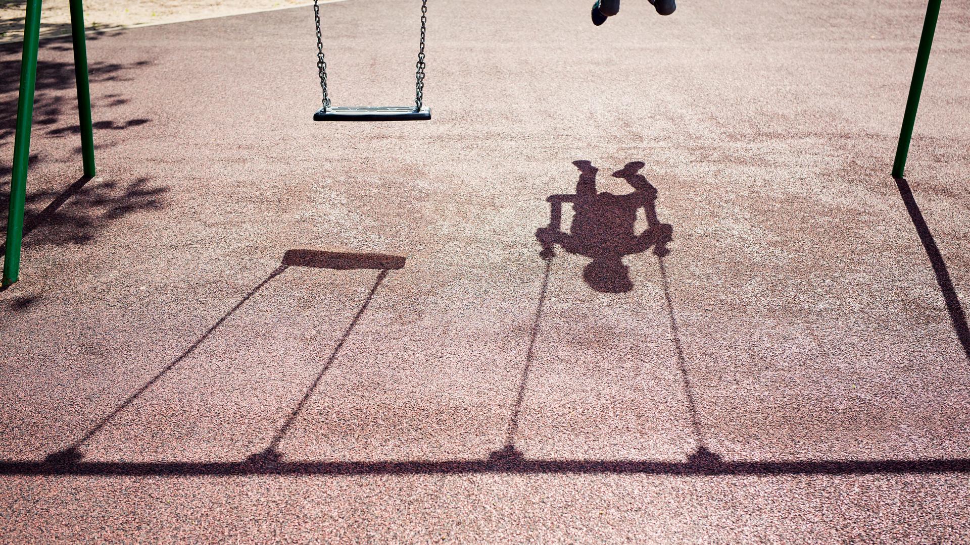 Der Schatten eines Kindes auf einer Schaukel ist auf dem Betonboden eines Spielplatzes zu sehen. Der Schatten eines Kindes auf einer Schaukel ist auf dem Betonboden eines Spielplatzes zu sehen.