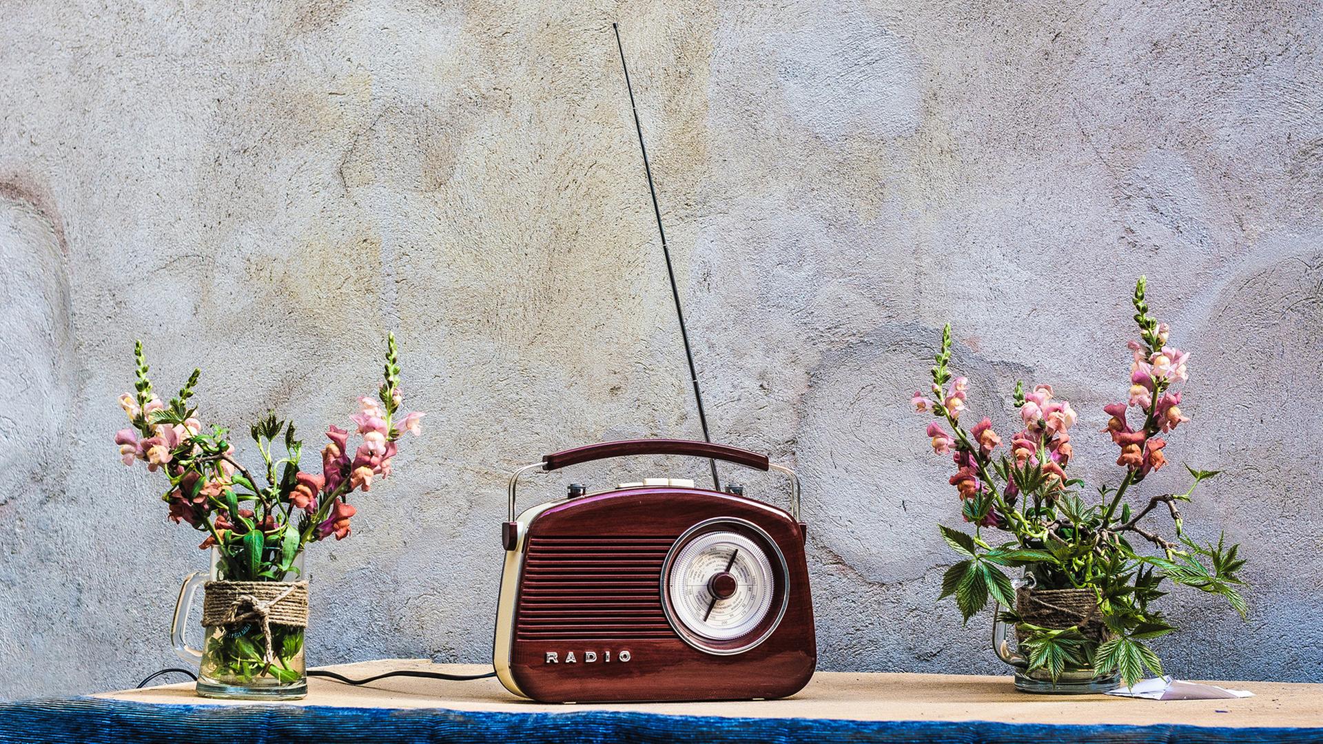 Auch wenn Radios längst nicht mehr so aussehen, fangen die Macher von „Angel Radio“ immer noch den Sound von Damals ein. Zu sehen: Ein rotbraunes, altes Handkofferradio mit ausgezogener Antenne in der Mitte eines Tisches. Daneben jeweils eine Blumenvase mit Löwenmäulchen. 