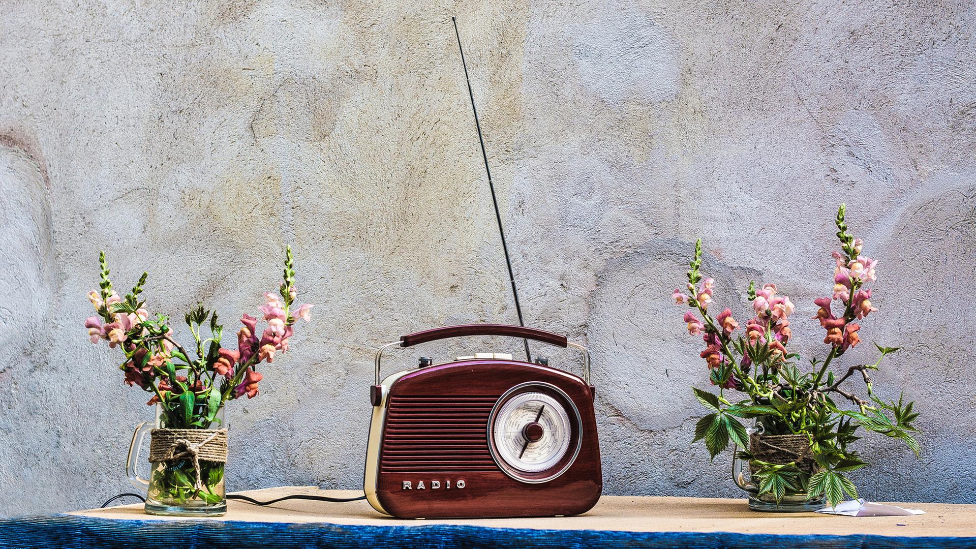 Auch wenn Radios längst nicht mehr so aussehen, fangen die Macher von „Angel Radio“ immer noch den Sound von Damals ein. Zu sehen: Ein rotbraunes, altes Handkofferradio mit ausgezogener Antenne in der Mitte eines Tisches. Daneben jeweils eine Blumenvase mit Löwenmäulchen. Auch wenn Radios längst nicht mehr so aussehen, fangen die Macher von „Angel Radio“ immer noch den Sound von Damals ein. Zu sehen: Ein rotbraunes, altes Handkofferradio mit ausgezogener Antenne in der Mitte eines Tisches. Daneben jeweils eine Blumenvase mit Löwenmäulchen.