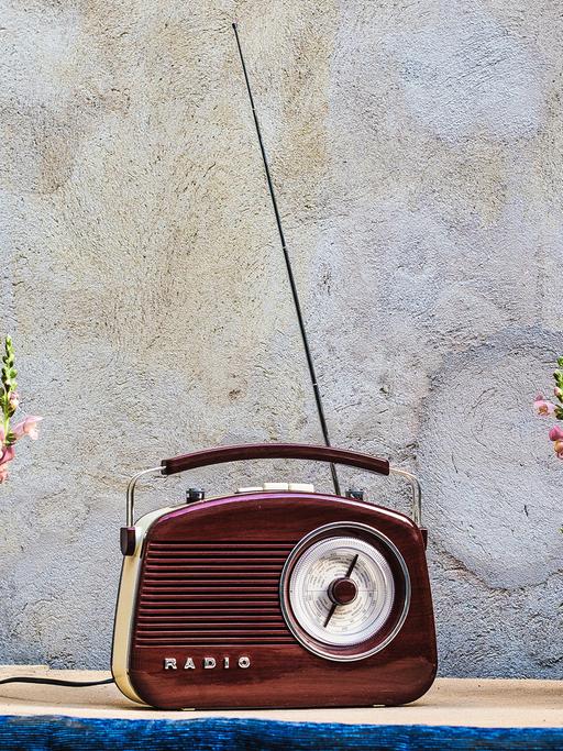 Auch wenn Radios längst nicht mehr so aussehen, fangen die Macher von „Angel Radio“ immer noch den Sound von Damals ein. Zu sehen: Ein rotbraunes, altes Handkofferradio mit ausgezogener Antenne in der Mitte eines Tisches. Daneben jeweils eine Blumenvase mit Löwenmäulchen. 