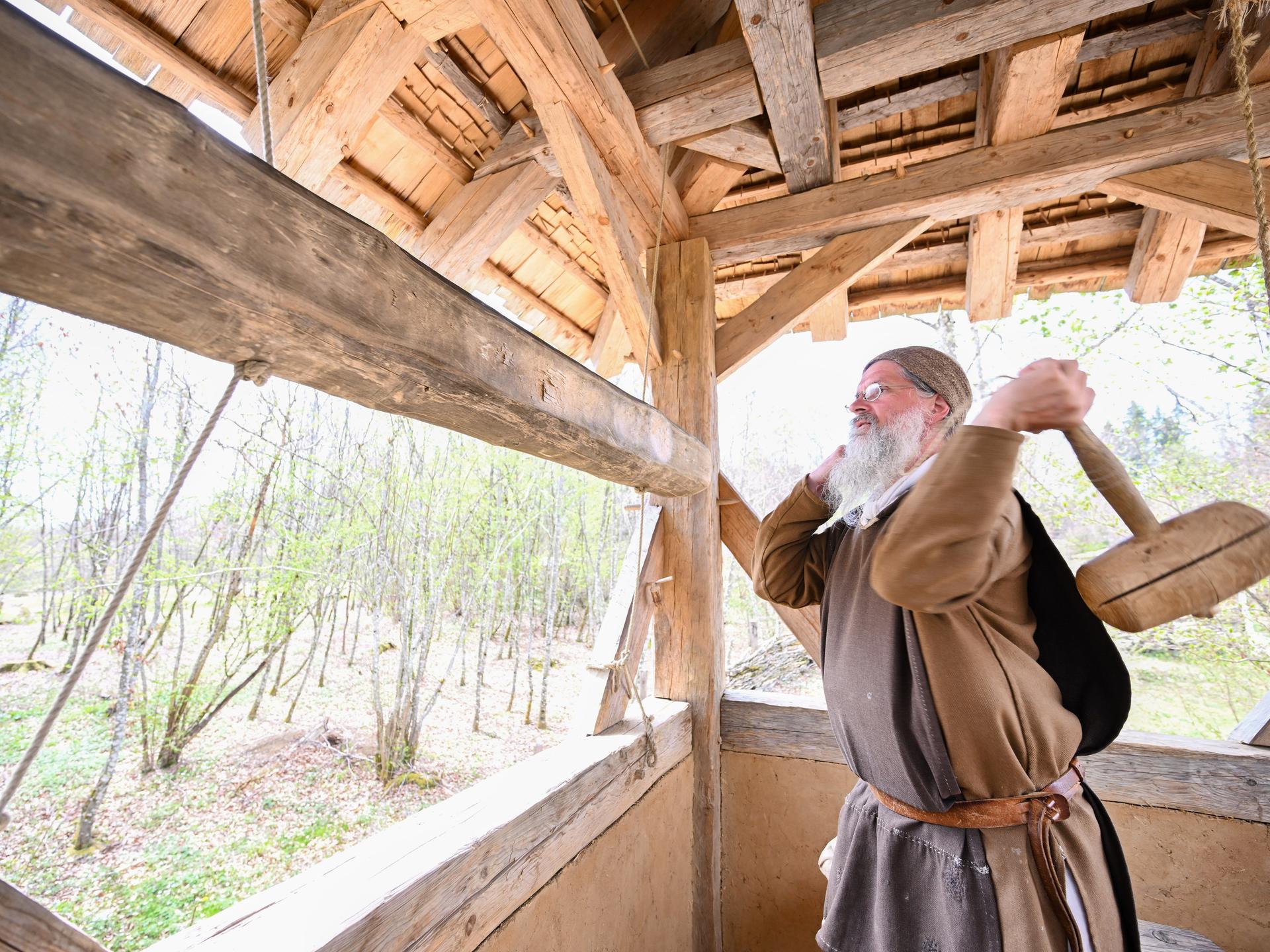 Ein Mann mit weißem Vollbart und in brauner Kutte steht auf einem Holzturm und schwingt einen Holzhammer. 