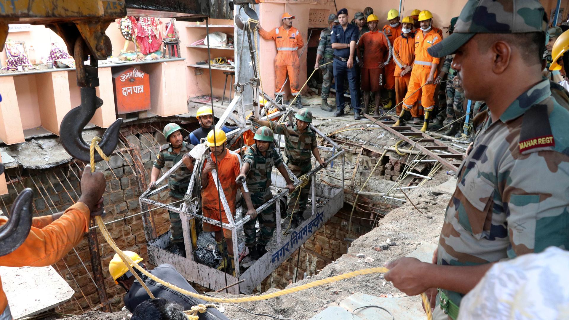 Das Bild zeigt indische Rettungskräfte, die mit einem Kran in den Brunnen unter dem Tempel gelassen werden. Weitere Rettungskräfte sehen dabei zu.