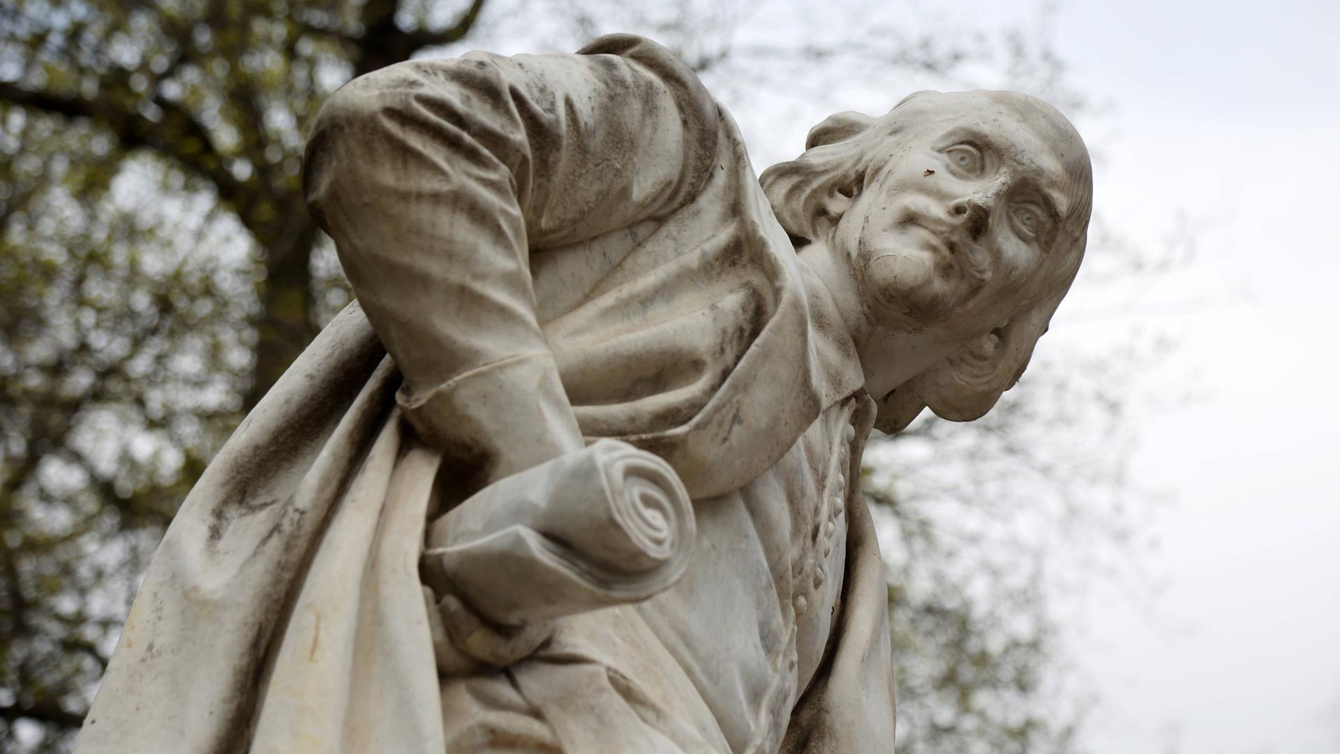 Weißes William Shakespeare Denkmal im Park an der Ilm in Weimar, auf dem die Figur mit einer Skriptrolle in der rechten Hand des angewinkelten Arms nach unten in die Kamera blickt.