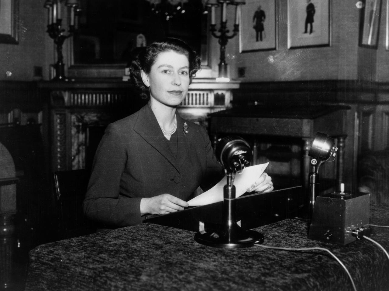 Elizabeth II. sitzt an einem Schreibtisch und spricht in Mikrofone.