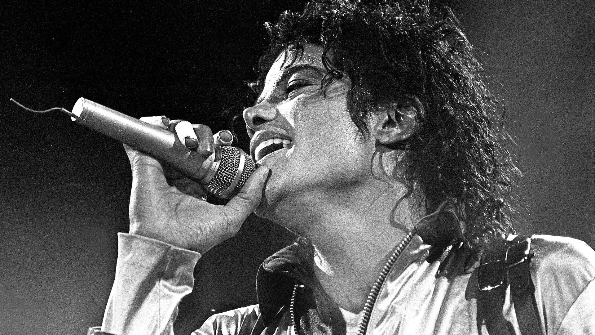 Schwarzweiße Nahaufnahme von Michael Jackson, der begeistert in ein Mikrofon singt. 