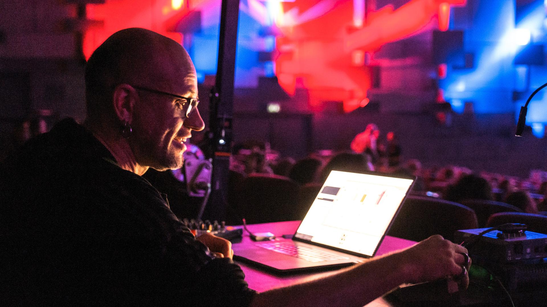 Der Medienkünstler Robert Henke schaut konzentriert auf einen Laptop und dreht an Reglern, der ganze Raum ist in rotes und blaues Licht getaucht. Der Medienkünstler Robert Henke schaut konzentriert auf einen Laptop und dreht an Reglern, der ganze Raum ist in rotes und blaues Licht getaucht.