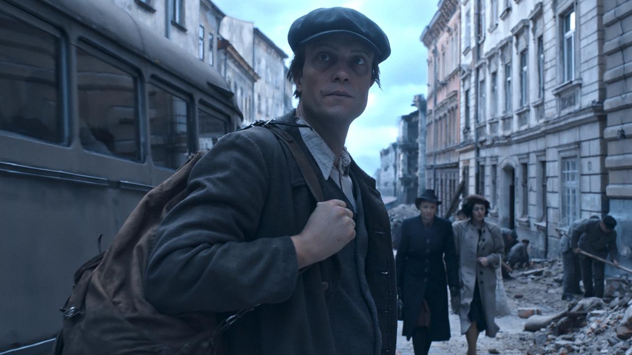 Eine Straßenszene in einer zerstörten deutschen Stadt nach dem 2. Weltkrieg. Ein Mann mit Rucksack blickt in die Kamera, zwei Frauen gehen an ihm vorbbei, Männer mit Schaufeln räumen Schutt weg.