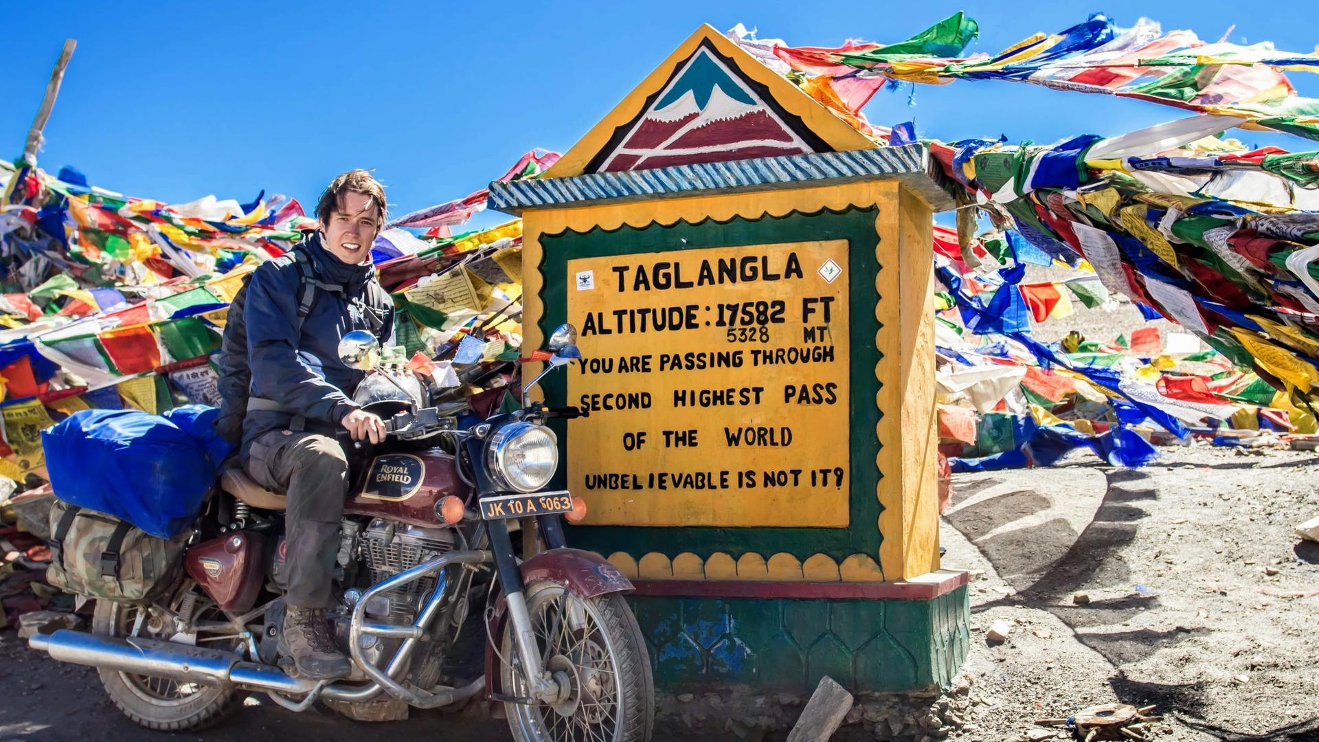 Erik Lorenz posiert auf einem Motorrad vor einem Schild, auf dem 'Taglangla' steht und an dem unzählige Schnüre mit bunten Gebetsfähnchen befestigt sind. Erik Lorenz posiert auf einem Motorrad vor einem Schild, auf dem 'Taglangla' steht und an dem unzählige Schnüre mit bunten Gebetsfähnchen befestigt sind.