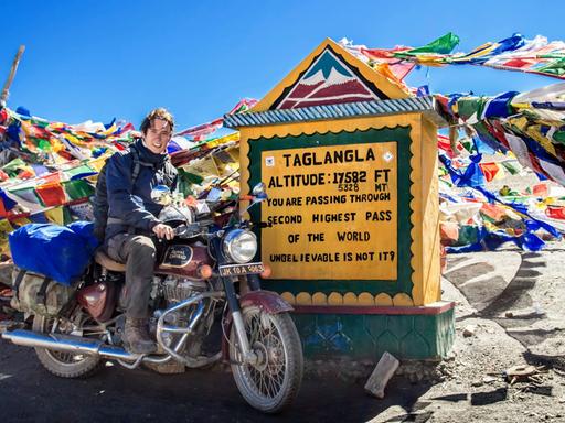 Erik Lorenz posiert auf einem Motorrad vor einem Schild, auf dem 'Taglangla' steht und an dem unzählige Schnüre mit bunten Gebetsfähnchen befestigt sind.