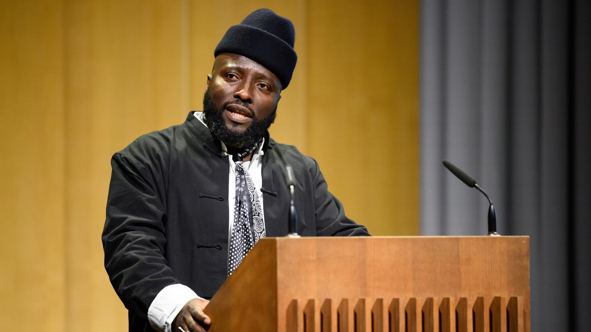 Bonaventure Soh Bejeng Ndikung spricht im Haus der Kulturen der Welt in Berlin an einem Pult vor einem Mikrofon. Bonaventure Soh Bejeng Ndikung spricht im Haus der Kulturen der Welt in Berlin an einem Pult vor einem Mikrofon.