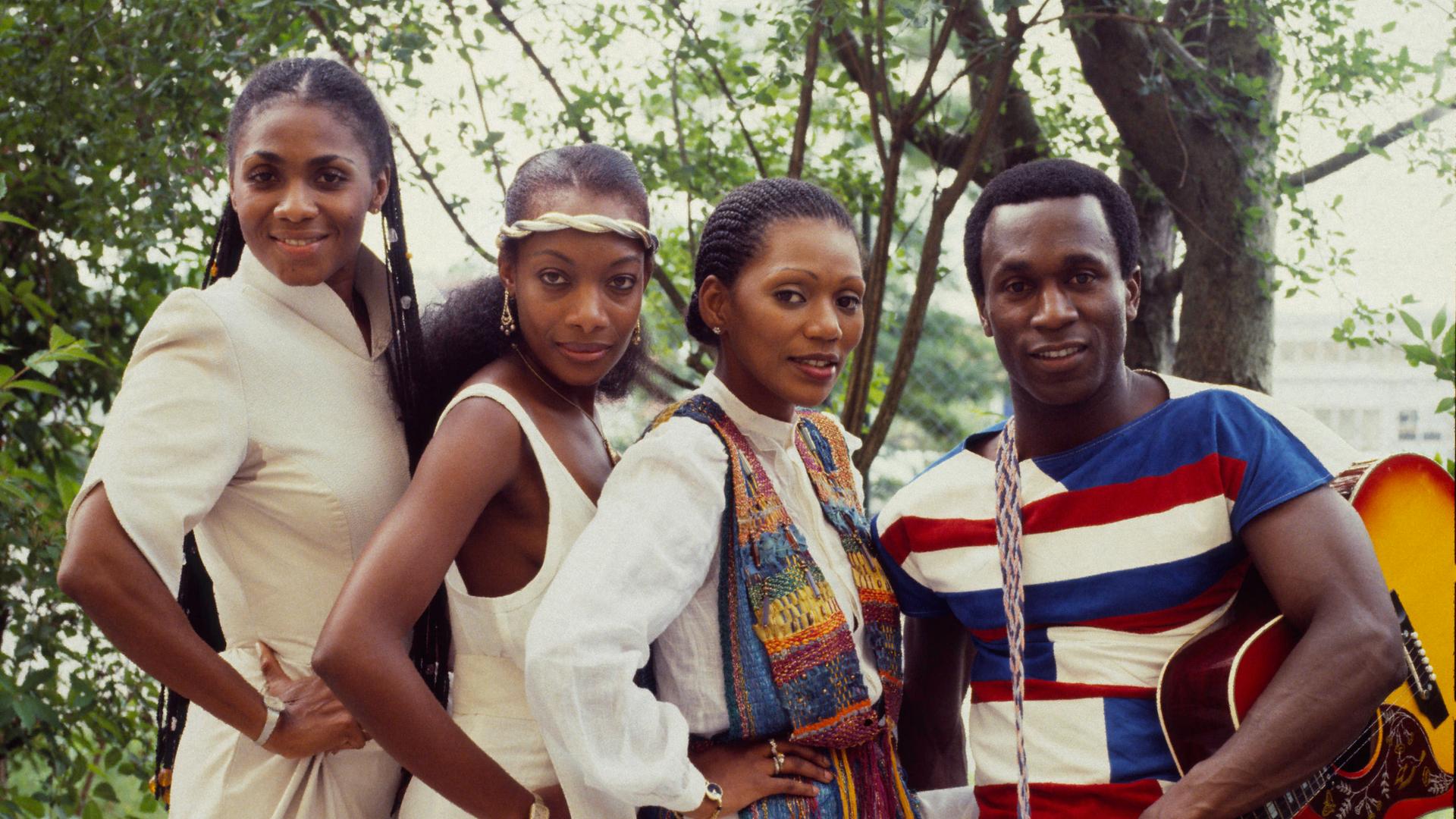 Die drei weiblichen und das männliche Mitglied der Band Boney M stehen dicht zusammen im Freien. Der Mann rechts hat eine Gitarre von der Schulter baumeln und hält sie mit einer Hand fest. Die drei weiblichen und das männliche Mitglied der Band Boney M stehen dicht zusammen im Freien. Der Mann rechts hat eine Gitarre von der Schulter baumeln und hält sie mit einer Hand fest.