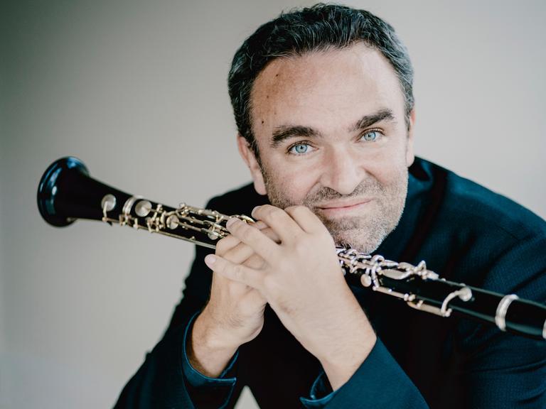 Der Klarinettist Jörg Widmann hält seine Klarinette vor sich, während er die Ellenbogen auf einem Tisch aufgestützt hat und schaut leicht lächelnd in die Kamera.