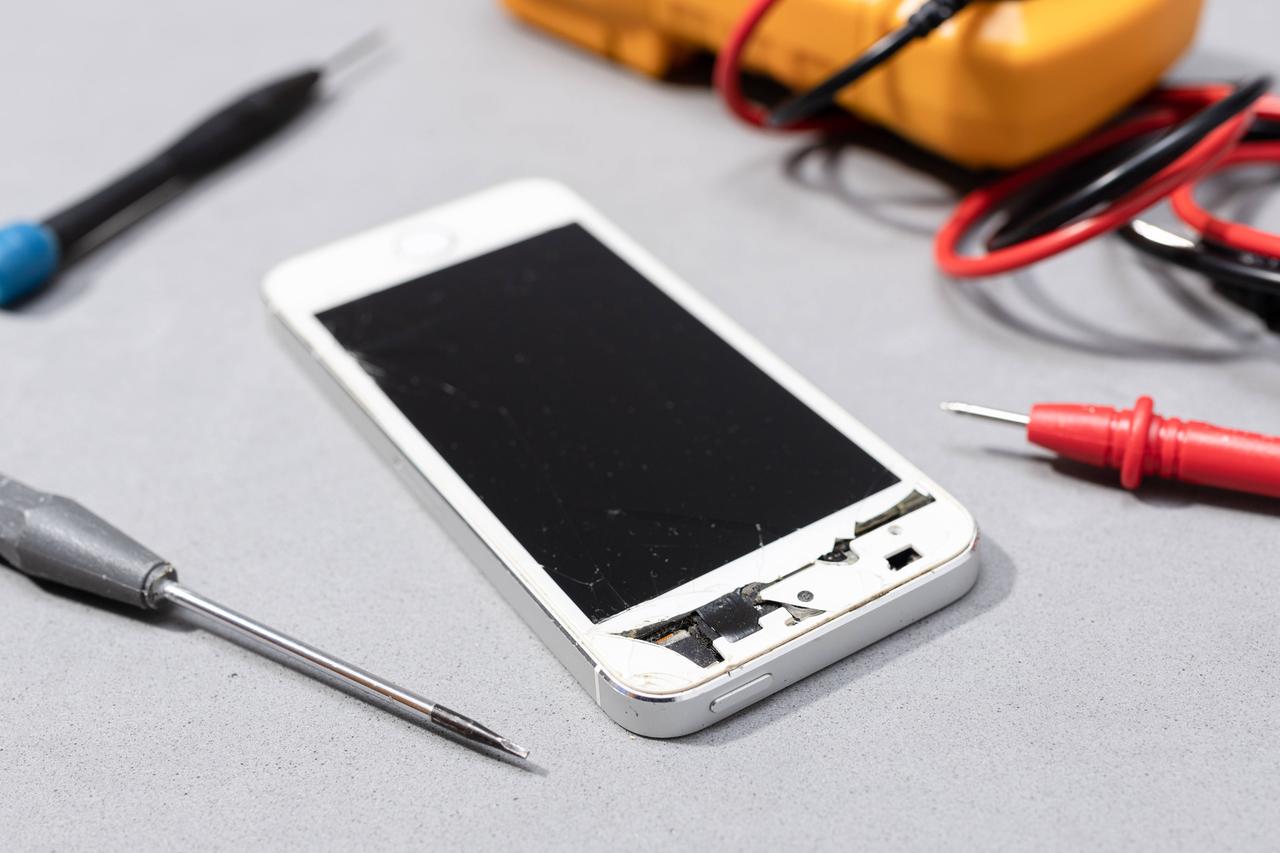 Reparatur von beschädigtem Handy: Zu sehen ist ein Smartphone mit kaputtem Bildschirm auf der Werkbank eines Elektronikgeschäfts.